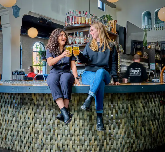 Jackie and Jessica sitting on the bar clinking beers together