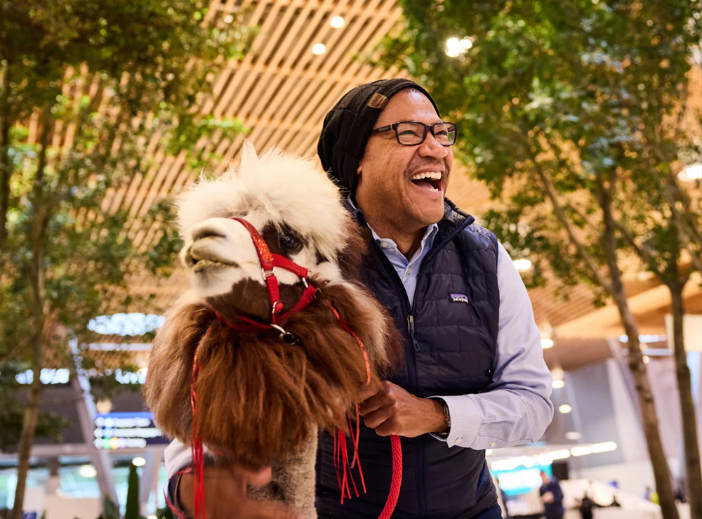 A man poses smiling next to a therapy llama with the mass timber roof shown behind him