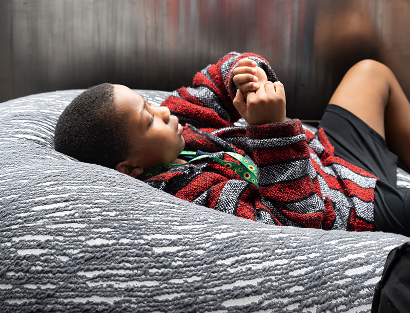 a boy lays on a beanbag chair in the Sendory Room wearing a sunflower lanyard while rubbing his hands
