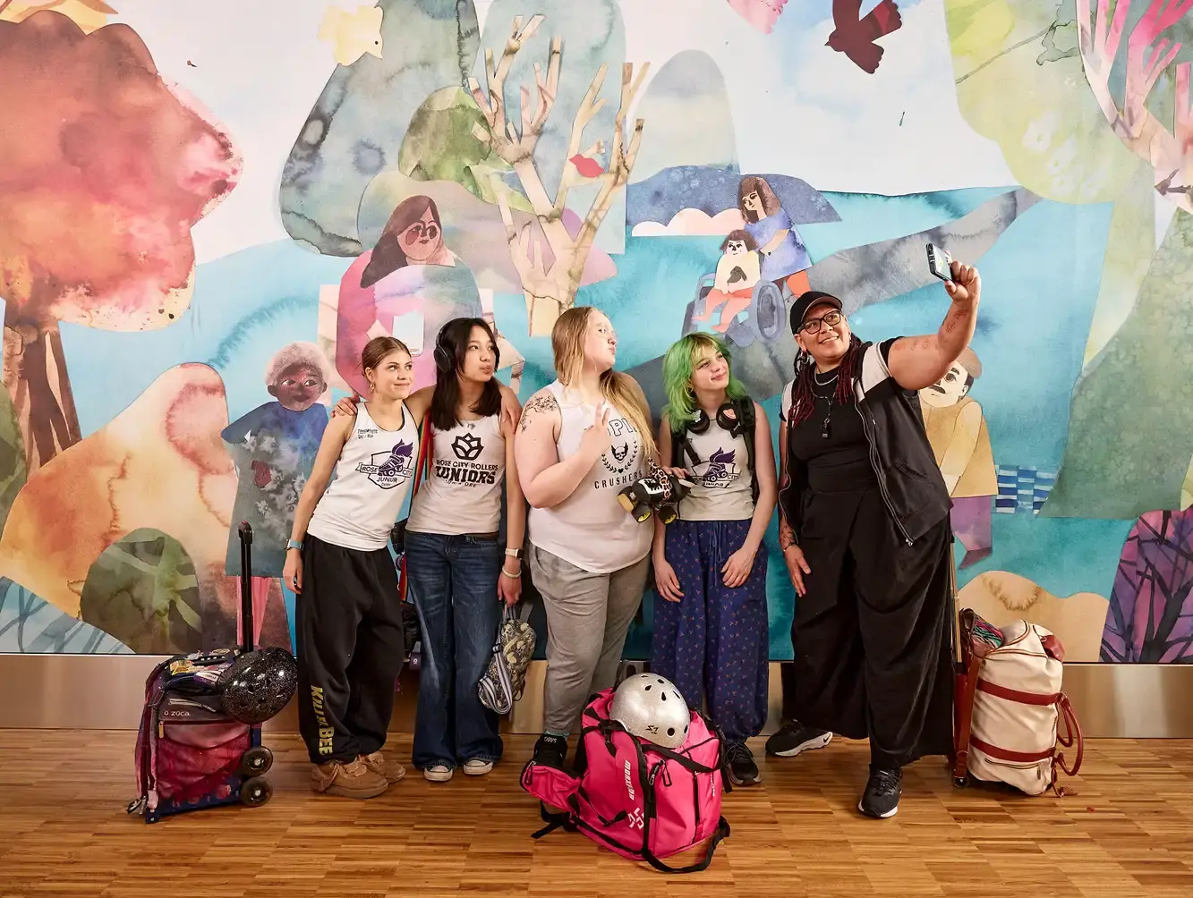 A Black adult woman holds a phone to take selfie with teenagers in front of mural