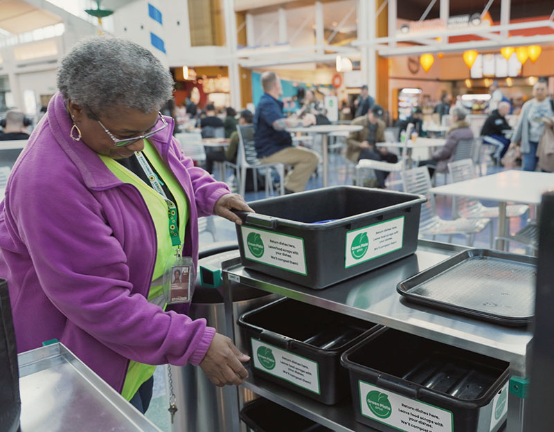 a Black woman is in the food court of concourse C supporting the bins which are used for the Green Plate Program