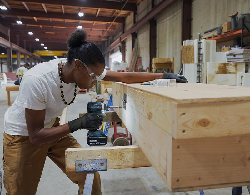a young Black woman wearing safety goggles drills a hinge into a wood frame in a warehouse