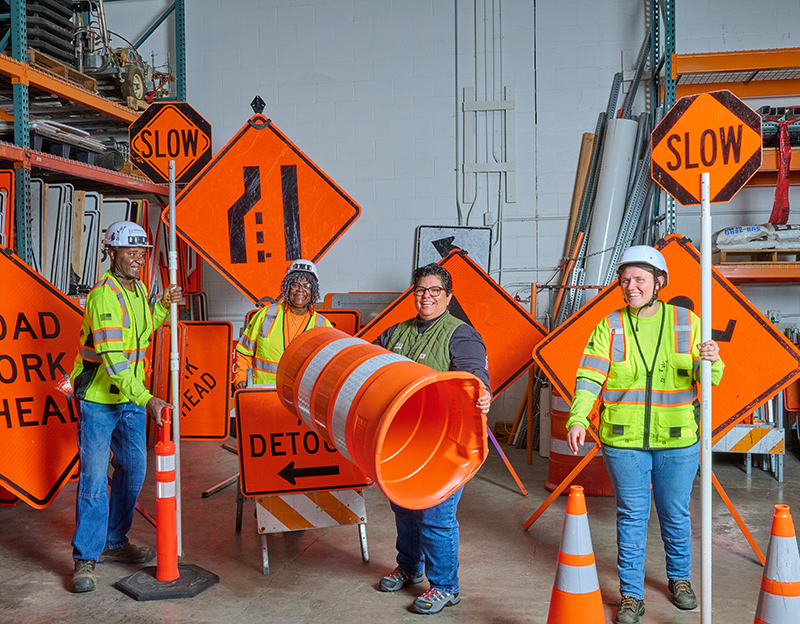 a diverse team of flagging professionals playfully pose with various bright orange road signs and cones