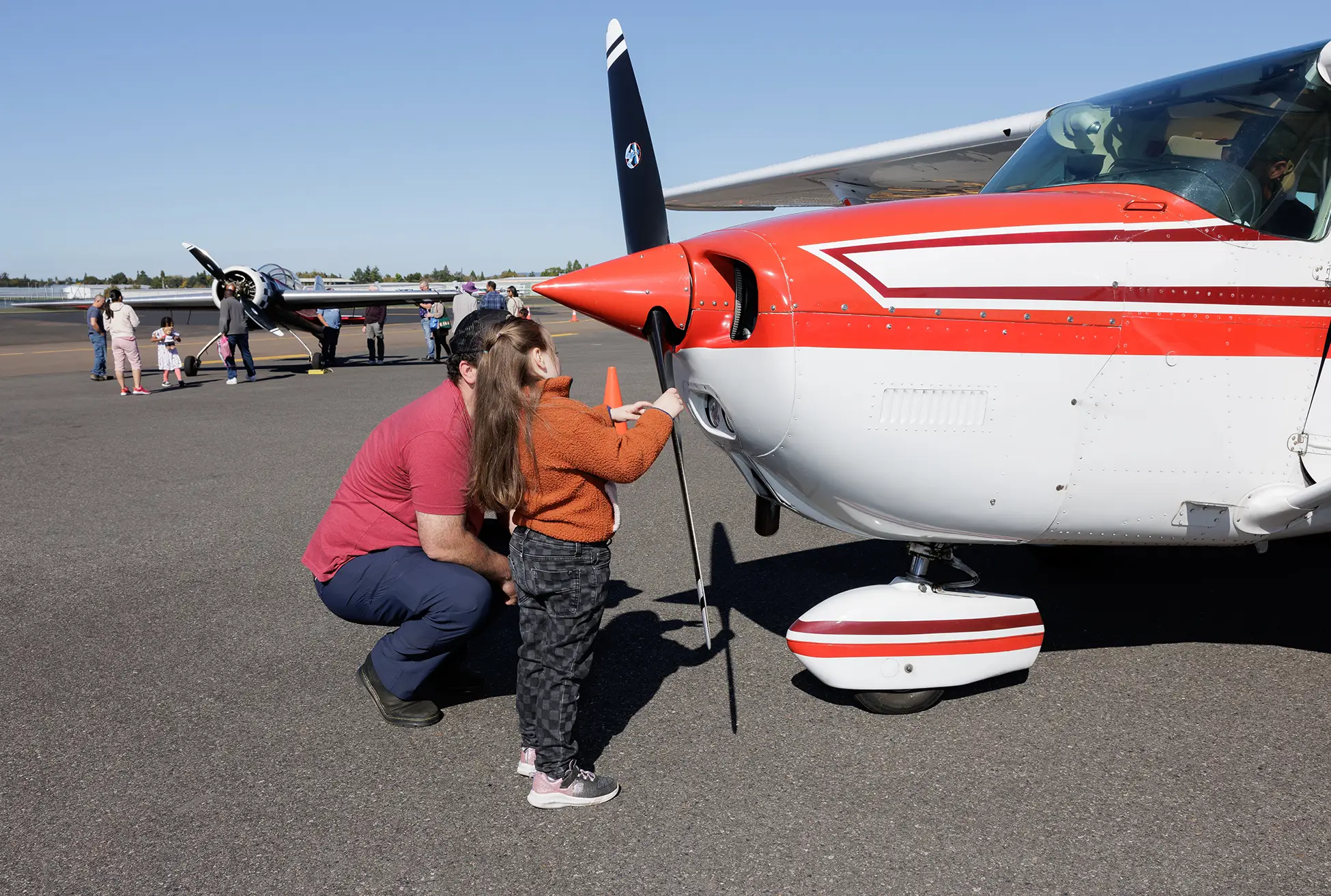 a father crouches down next to his daughter who is inspecting the propellor of a small plane during the Hillsboro Airport Block Party
