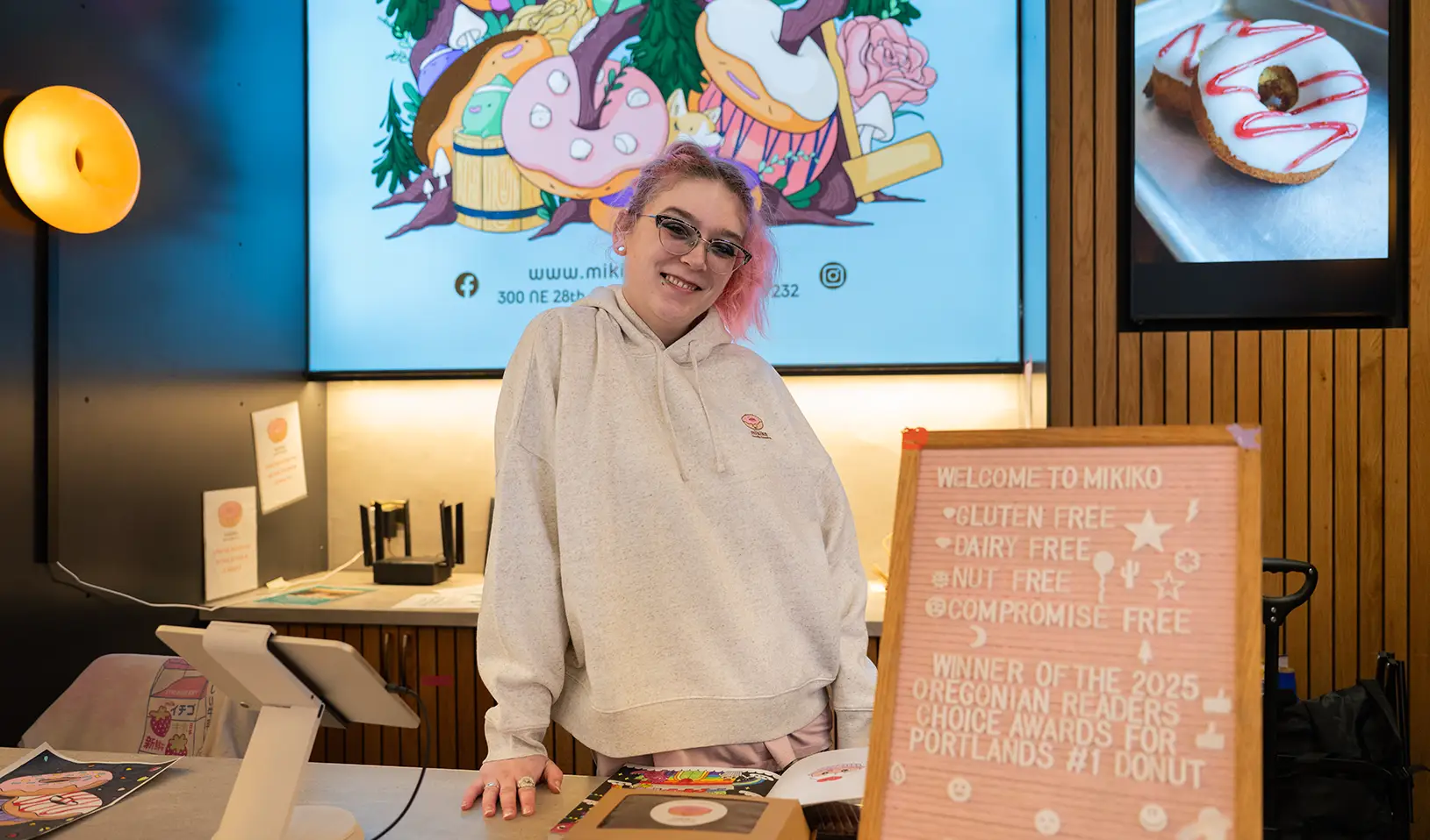 A white woman with pink hair is standing in front of an illuminated sign and a digital display monitor wearing a branded hoodie
