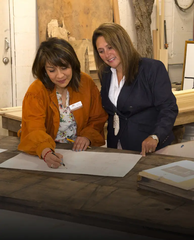 a Latina woman draws on an architectural plan while a white woman looks on both smiling