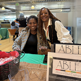 two Black women are smiling while tabling during an ERG fair for the ABLE group