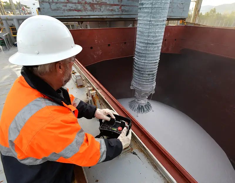 A man in a safety jacket and hard hat uses a controller to operate drybulk loading and unloading equipment