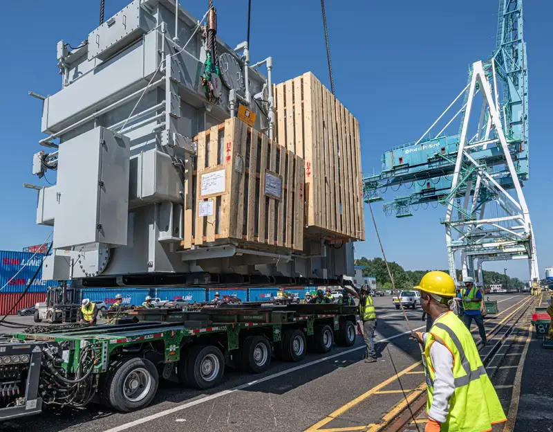 A man in a safety vest and hardhat holds a rope attached to a transformer being unloaded from a vessel at Terminal 6