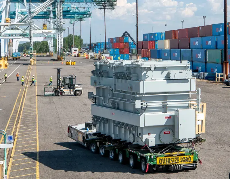 A man in a safety vest and hardhat holds a rope attached to a transformer being unloaded from a vessel at Terminal 6