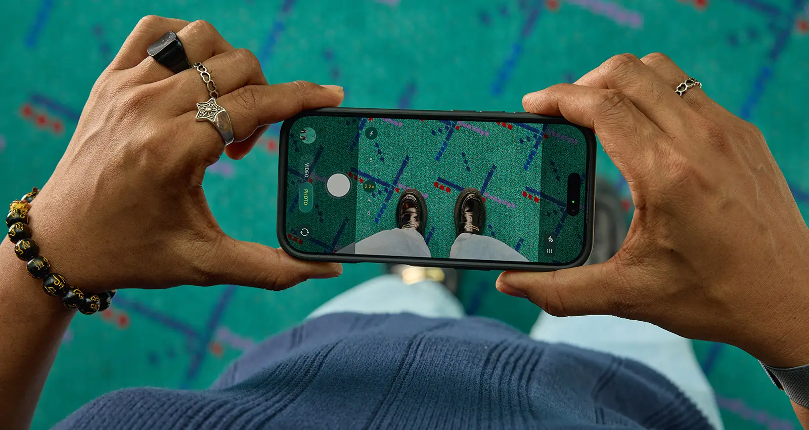 a dark-skinned man's hands taking a shoe selfie with their smartphone wearing various rings and a bracelet