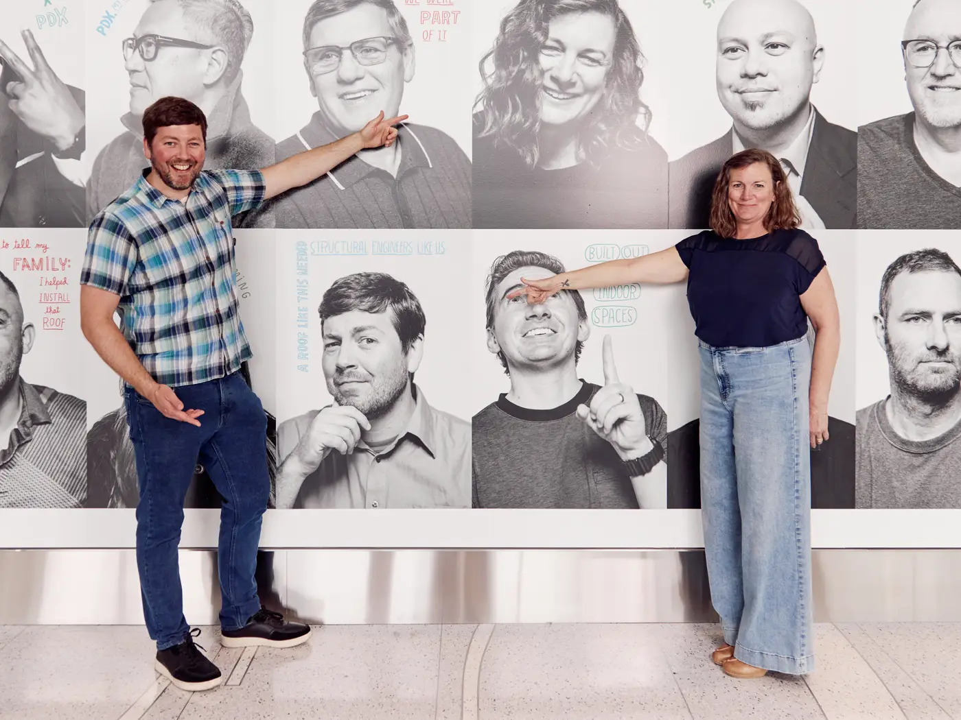 a man and woman pose in front of their photos within the mural, pointing at the other person's photo