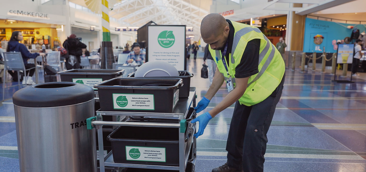 A Latino male employee is wearing a safety vest and gloves while removing the used dishes from the dining area