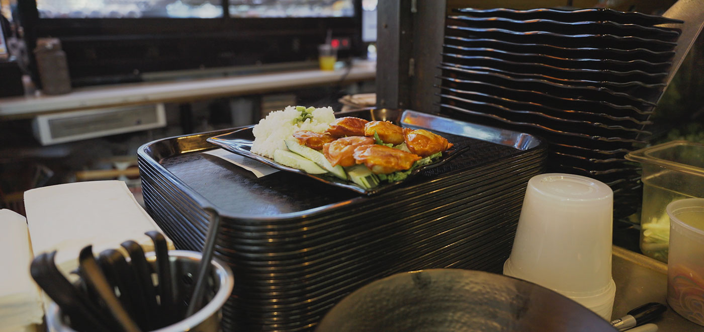 A chicken dish from Bambuza sits on top of a reusable plate on top of a stack of plastic trays in the serving area