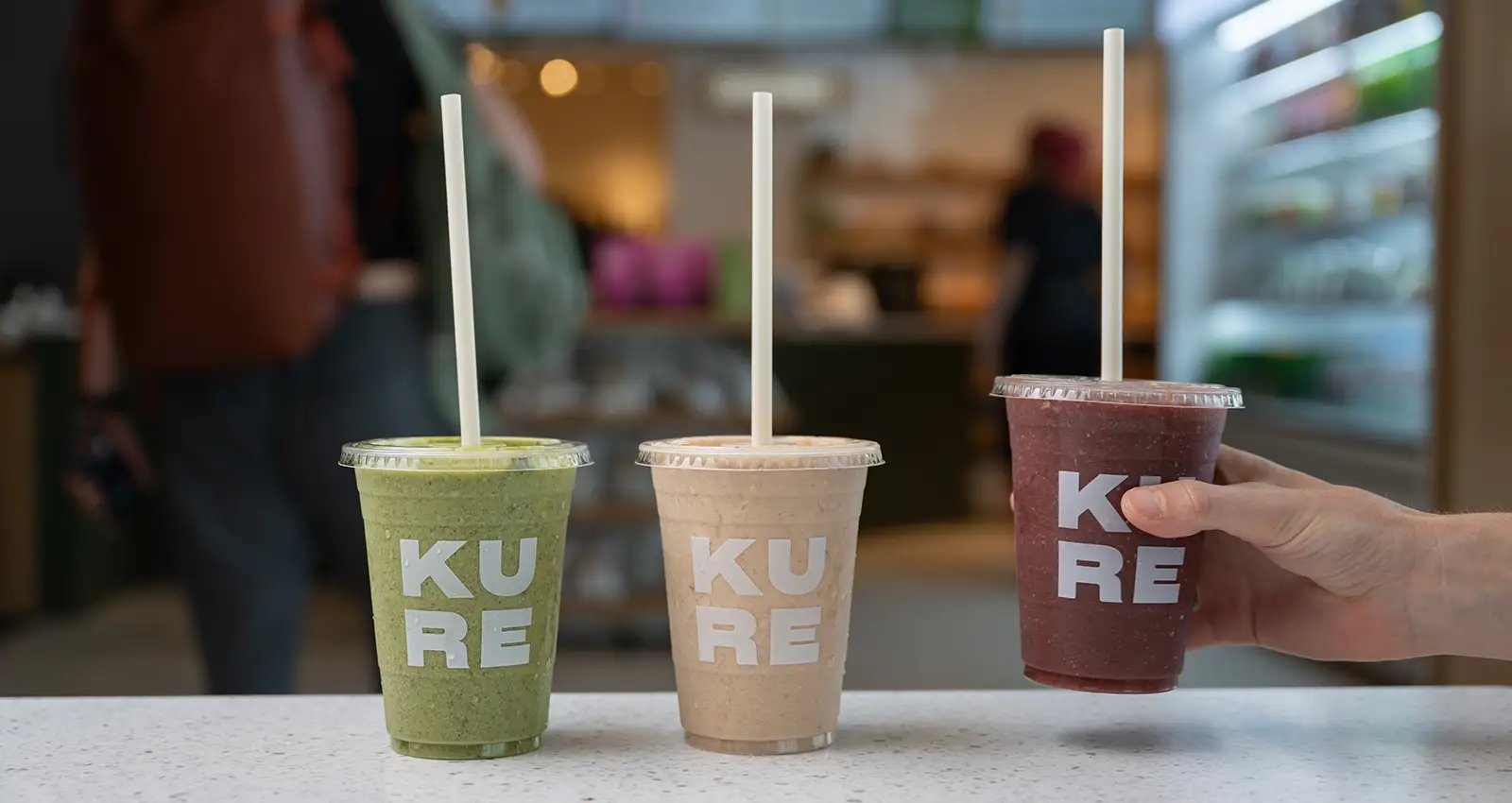 three Kure smoothies, one green, one beige, and one berry-colored with a man's hand grabbing one